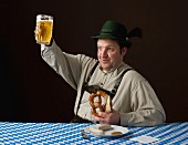 A stereotypical German man wearing lederhosen and eating a pretzel and white sausage with a beer