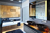 Modernes Bad, Waschtisch mit zwei schwarzen Aufbaubecken, oberhalb Spiegel mit integrierter Beleuchtung, im Hintergrund Badewannenfront und Wand mit Fliesen in Messing-Optik