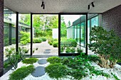 Indoor garden in vestibule with ground-cover plants and tree amongst white pebbles and view of garden through glass facade