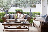 Sofas with wicker frames and scatter cushions around simple wooden coffee table on terrace; low clipped hedge in background