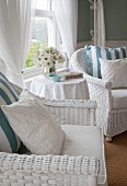 White-painted wicker armchairs with cushions and vase of white flowers on round side table in front of window