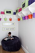 Little boy sitting on giant bean bag in corner below colourful metal buckets hung below white bracket shelves