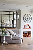 Silver-painted bistro table next to pale chaise, framed mirror on wall, stacked firewood in fireplace and antique wall clock with Roman numerals