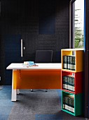 Bunte Holzmodule mit Büchern übereinander gestapelt, dahinter Schreibtisch mit orangefarbener Kunststoffplatten