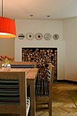 Upholstered chairs around wooden table below pendant lamps with orange lampshade; stacked firewood in fireplace in background