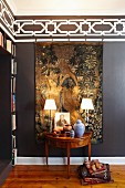 Table lamps on Biedermeier-style console table in front of antique Flemish tapestry on dark brown wall with stencilled black and white frieze