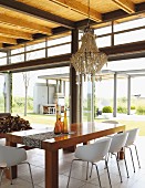 Chandelier above dining set with elegant wooden table and shell chairs; view into garden through steel and glass facade