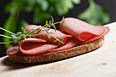A slice of bread topped with salami and fresh oregano on a wooden table in a garden