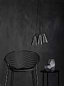 Wire mesh chair and matte grey vase on side table below bundle of shiny, metallic lampshades against black wall