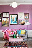 Polstertisch und geschwungene Couch mit bunten Kissen vor lila Tapete an Wand mit gerahmter Bildersammlung