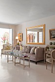Eleganter Wohnraum in hellen Farbtönen, antiker Couchtisch und Sofa mit drapierten Kissen, vor Goldrahmenspiegel an Wand