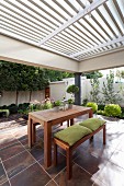 Wooden table and benches with green seat cushions on tiled terrace below pergola with white, wooden, slatted sunshade