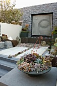 Meditation garden; bowl of succulents on stone plinth, benches in raised area and tall stone wall with picture of Buddha in niche