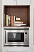 Microwave in kitchen cabinet below cookery books in wooden shelf compartment