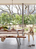 Modern table and bench set on pale grey concrete floor in front of glass wall with view of garden