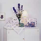 Schachteln mit lavendelfarbenem Toile de Jouy Muster und blaue Glasflaschen mit Rittersporn auf weisser Kommode