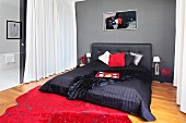Schwarzes Doppelbett mit gepolstertem Kopfteil und Tagesdecke vor grauer Wand; auf Boden roter Teppich mit Sprenkelungen