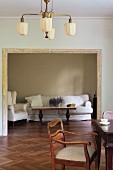 Upholstered wooden chair at table below ceiling lamp in Biedermeier style; wide doorway in background with view of coffee table and white sofa