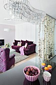 An organically shaped crystal chandelier above a the reflective surface of a kitchen bar with elegant, purple upholstered furniture in the background