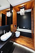 An elegant bathroom with a black and white bathtub and washstand, a wooden panelled toilet section and a modern lamp