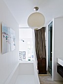 Modern bathtub below white spherical lamp in front of glass wall