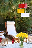 Pendant lamps upcycled from cake-tins painted various colours above terrace table, wooden lounger with white cushions in front of palm tree