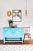 Pale blue, carved dresser and country-house wooden chair below modern artwork on white-painted, wood-clad wall and retro pendant lamp