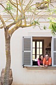 Ein Paar schaut aus Fenster, vor dem Haus ein Mimosenbaum und ein mediterranes Tongefäss