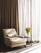 Pale armchair and vase of tulips on side table in front of floor-length curtains on window