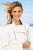 A young blonde woman on a beach wearing a white leather jacket