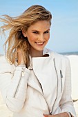 A young blonde woman on a beach wearing a white leather jacket