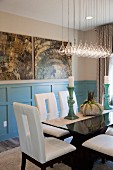 Elegantes Esszimmer mit weissen Stühlen, Esstisch aus Glas, Designer-Hängeleuchte & abstrakten Gemälden über blauer Wandverkleidung aus Holz
