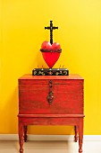 Kruzifix über rotem Herz auf Schränkchen vor leuchtend gelber Wand