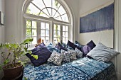 Bett mit blauem Bettüberwurf und blauen Zierkissen vor Rundbogenfenster