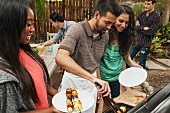 A man at a barbecue using tongs to give two women vegetable skewers