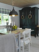 Schlichter Esstisch und weiße Küchenstühle unter Vintage Hängeleuchte, im Hintergrund dunkel lackierter Bauernschrank mit Kränzen an Türen