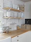 Küchenecke mit Geschirr auf Wandkonsolen an weisser Holzwand, darunter Aufbewahrungsgläser auf Arbeitsplatte