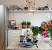 Table set with blue and white crockery and spring flowers; potted herbs on kitchen counter in background