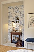 Table lamp, lantern and silver bowl on antique table in niche with floral wallpaper above fawn figurine on denim cushion