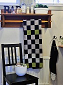 Chequered towel on towel rail with ornaments on wooden shelf above black-painted wooden chair