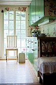 White chair in front of floor-to-ceiling lattice window and fitted cupboards with green doors in rustic kitchen