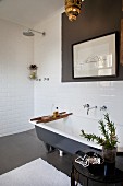 Plant on side table next to free-standing bathtub, framed drawing on wall and floor-level, open shower in corner of white-tiled bathroom