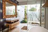 Partially visible bathtub in front of glazed shower area and view of terrace; washstand to one side below mirror with carved wooden frame on papered wall