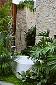 Free-standing bathtub with floor-mounted taps in idyllic garden in front of high stone wall with outdoor shower