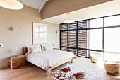 Kuhfell auf edlem Dielenboden vor schlichtem Doppelbett in grosszügigem Schlafzimmer, an Fensterfront Schiebeelemente aus Holzlamellen