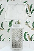 Table lamp with white ceramic base with ornate perforated pattern and translucent lampshade against fern-patterned wallpaper