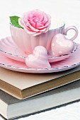 Rosafarbene Tasse auf Bücherstapel dekoriert mit Rosenblüte & Dekoherzen