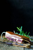 A slice of pancetta on a wooden board with rosemary and a knife