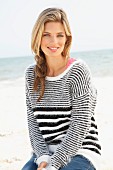 A young blonde woman sitting on a beach wearing a black-and-white striped knitted jumper