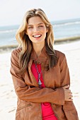 A young blonde woman on a beach wearing a brown leather jacket and a pink top